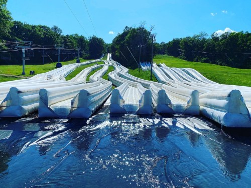 A series of inflatable water slides on a sunny day, surrounded by green hills and a clear blue sky.