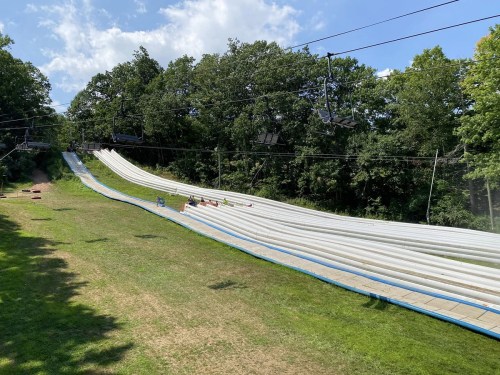 A summer slide with multiple lanes, surrounded by trees and a clear blue sky, with a ski lift overhead.