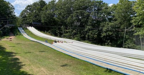 A summer scene of a water slide with people sliding down, surrounded by green trees and a clear blue sky.