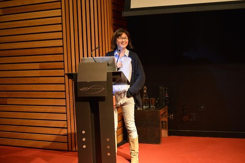A woman stands at a podium, smiling and speaking to an audience in a modern conference setting.