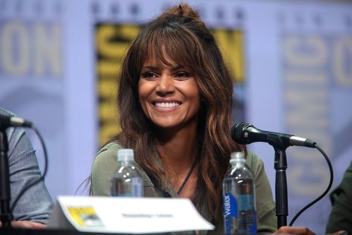 A smiling woman with long, wavy hair sits at a panel table with a microphone and water bottles in front of her.