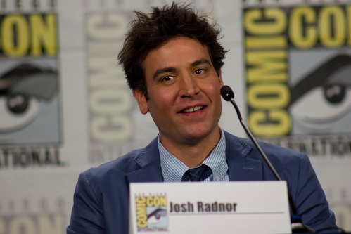 A man with tousled hair smiles at a panel, sitting behind a nameplate that reads "Josh Radnor" at Comic-Con.