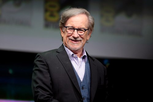 A smiling man with glasses and gray hair, wearing a black suit and blue vest, stands on stage at an event.