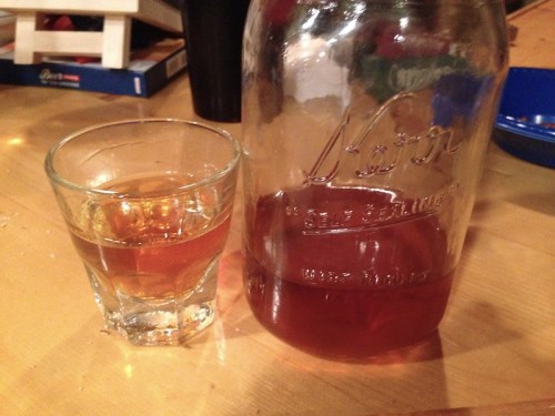 A glass of amber liquid next to a large jar filled with the same liquid on a wooden table.
