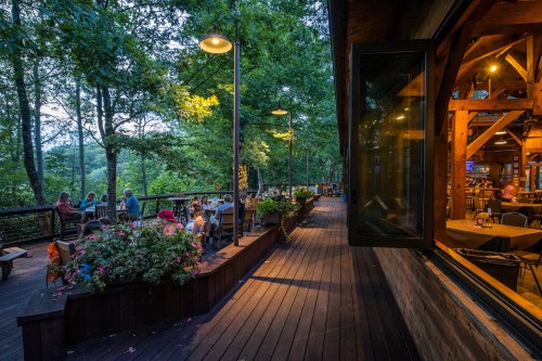 A cozy restaurant deck surrounded by trees, with diners enjoying the evening ambiance and warm lighting.
