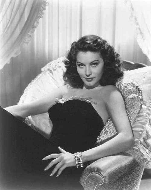 A glamorous woman in a black dress lounges on a sofa, exuding elegance with wavy hair and a sparkling bracelet.