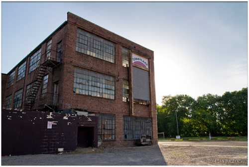 Abandoned brick building with large windows, a faded sign, and a fire escape, set against a bright sky.