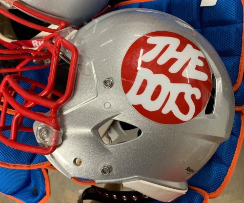A silver football helmet with a red logo that reads "THE DOTS" on the side, resting on blue padding.
