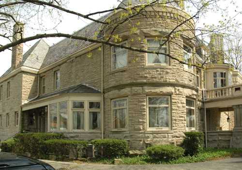 A large stone house with a rounded turret, surrounded by greenery and trees, showcasing intricate architectural details.