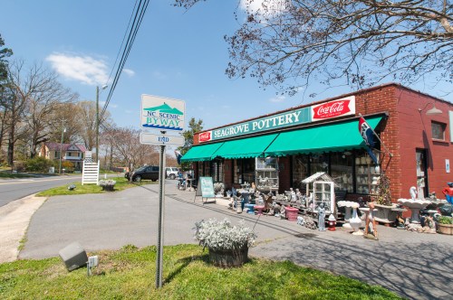 A pottery shop with a green awning, displaying various pottery items, next to a scenic byway sign on a sunny day.