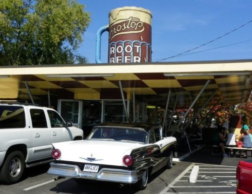 A vintage car parked outside a retro root beer stand with a large root beer mug sign overhead.