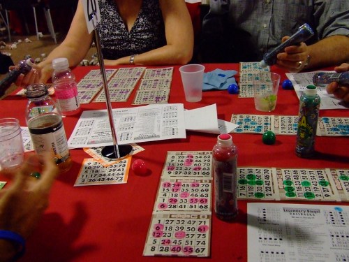 A bingo game in progress with players' hands, cards, and drinks on a red table.