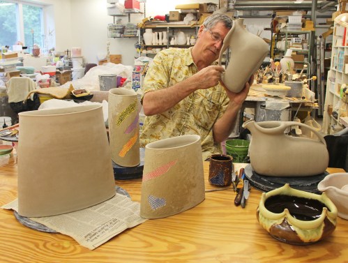 A potter shapes clay pieces in a studio, surrounded by tools and finished pottery on a wooden table.