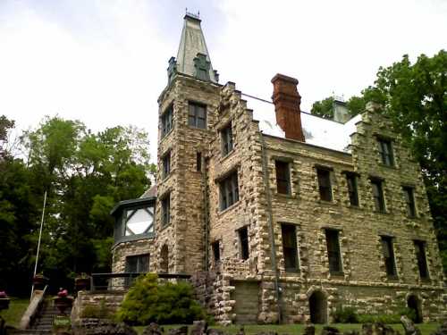 A historic stone castle with a tall spire, surrounded by trees and greenery, showcasing intricate architectural details.