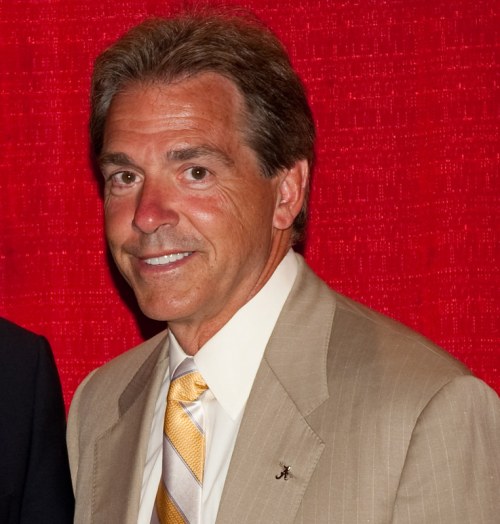 A man in a light suit and yellow tie smiles against a red backdrop.