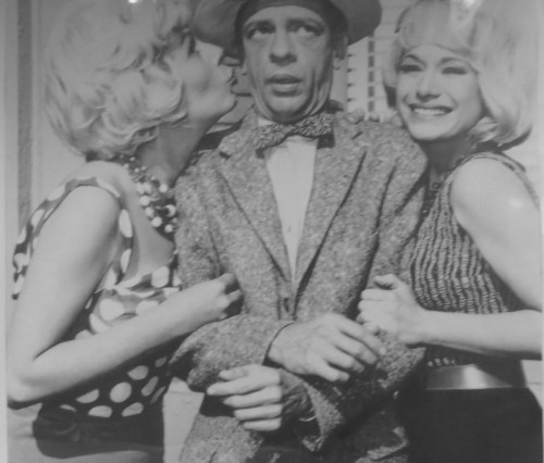 Two women in polka dot outfits kiss a man in a suit, all smiling in a playful, vintage black-and-white photo.