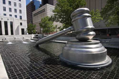 A large metallic gavel sculpture partially submerged in a water feature, surrounded by trees and buildings.