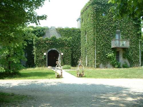 A stone building covered in ivy, featuring an arched entrance and a balcony, surrounded by greenery and a gravel path.