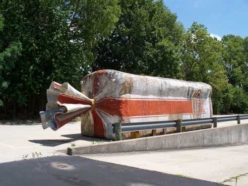 A large, weathered concrete structure resembling a giant arrow, painted with red and white stripes, set in a green park.