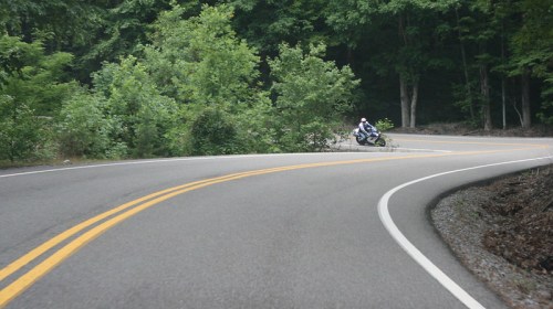 A motorcycle leans into a curve on a winding road surrounded by lush green trees.