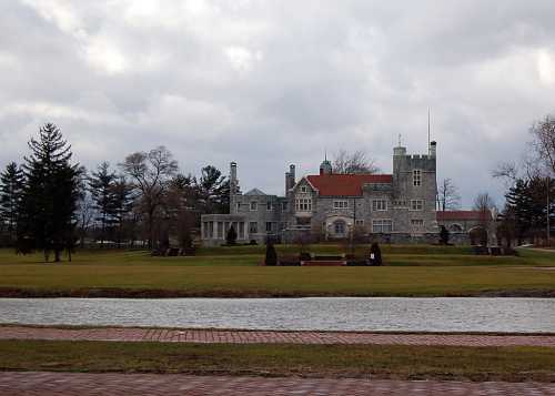 A large stone mansion with a red roof, surrounded by green lawns and trees, under a cloudy sky.