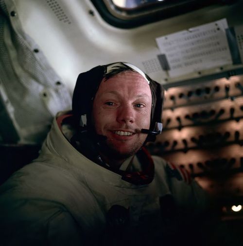 Astronaut in a space suit smiling inside a spacecraft, with control panels visible in the background.