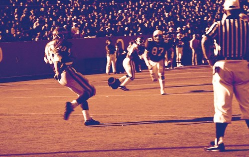 A football player kicks the ball on a field, with teammates and a referee in the background, during a game.