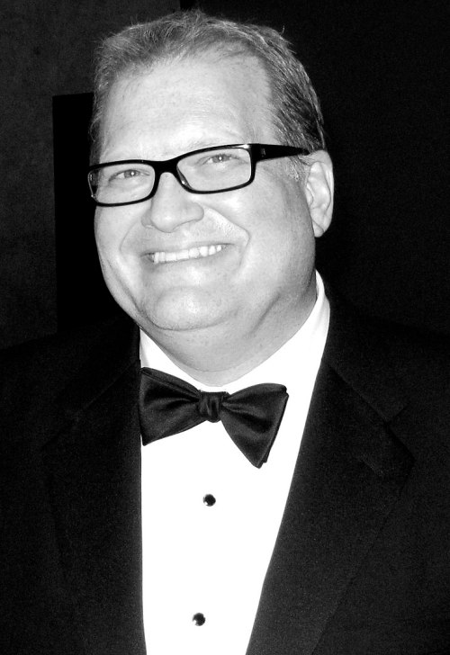 A smiling man in a tuxedo and bow tie, wearing glasses, poses for a photo in black and white.