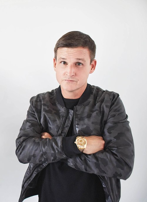 A man in a black camo jacket stands with arms crossed, wearing a gold watch and a serious expression against a light background.