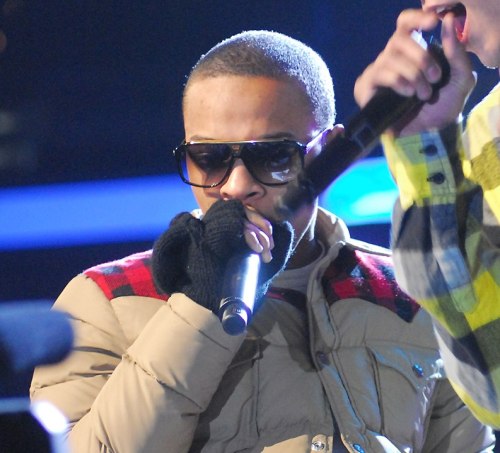 A performer wearing sunglasses and a puffy jacket holds a microphone, singing on stage.