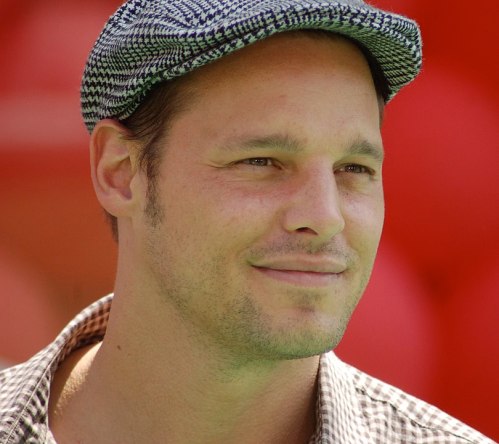 A man wearing a checkered shirt and a patterned cap smiles, with a blurred red background.
