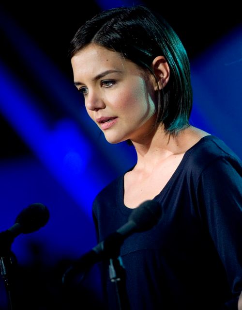 A woman with short hair stands at a microphone, looking down thoughtfully against a blue-lit background.