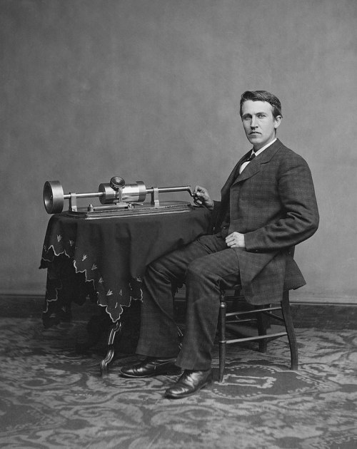 A man in a suit sits at a table with an early phonograph, posing for a portrait in a vintage setting.