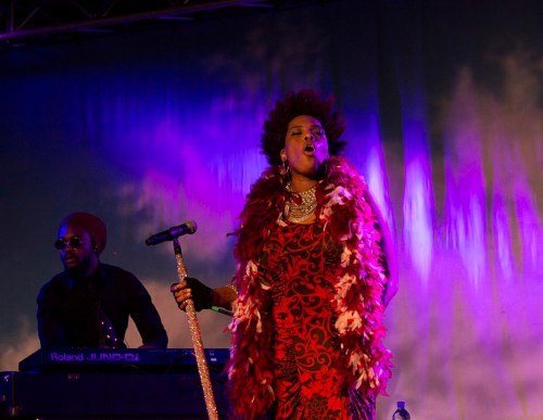 A singer in a red dress and feather boa performs on stage, with a keyboardist in the background under colorful lights.
