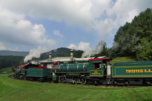 Two vintage steam locomotives curve around a green hillside, with smoke billowing against a backdrop of trees and clouds.
