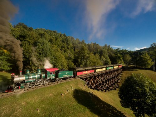 A vintage steam train crosses a wooden trestle bridge surrounded by lush green trees under a blue sky.