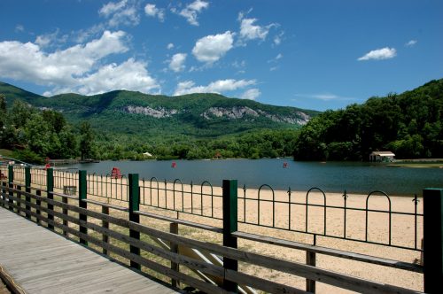 A serene lake surrounded by green mountains under a blue sky, with a sandy beach and wooden walkway in the foreground.
