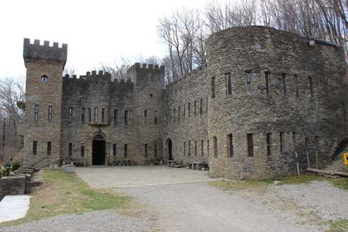 A stone castle with towers and arched windows, surrounded by trees and a gravel path.