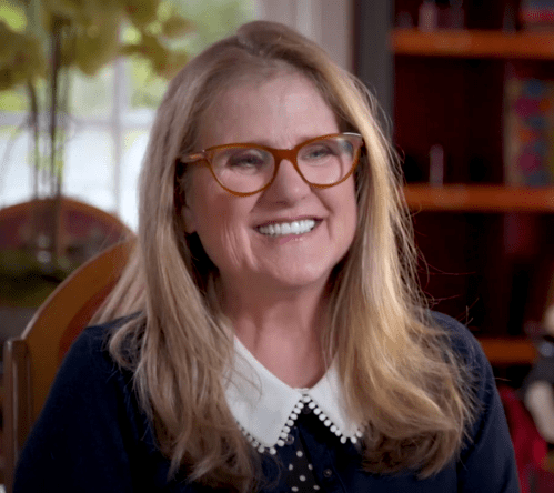 A smiling woman with long hair and glasses sits in a cozy room, wearing a navy sweater with a white collar.