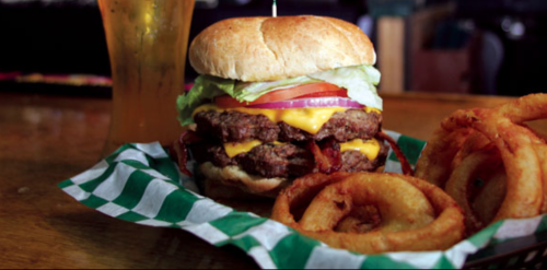 A juicy cheeseburger with lettuce, tomato, and onion, served with crispy onion rings and a drink in the background.