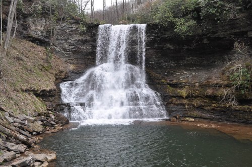 A serene waterfall cascading over rocky cliffs into a calm pool, surrounded by trees and natural greenery.