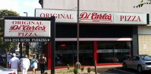 Exterior of Di Carlo's Pizza, featuring a sign with the restaurant's name and a parking sign, with people walking by.