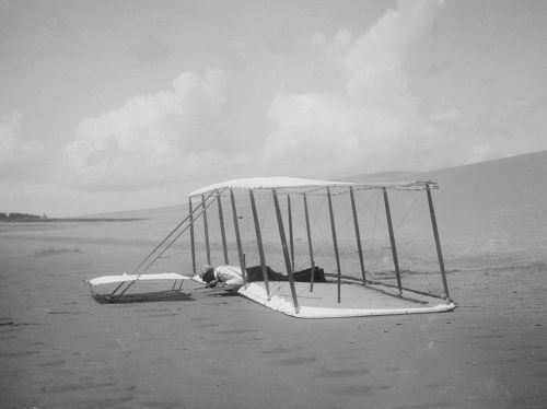 A historic black-and-white photo of a man lying on a sand dune beside a primitive glider.