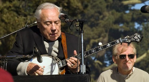 A musician plays the banjo on stage, while another man smiles in the background, surrounded by trees.