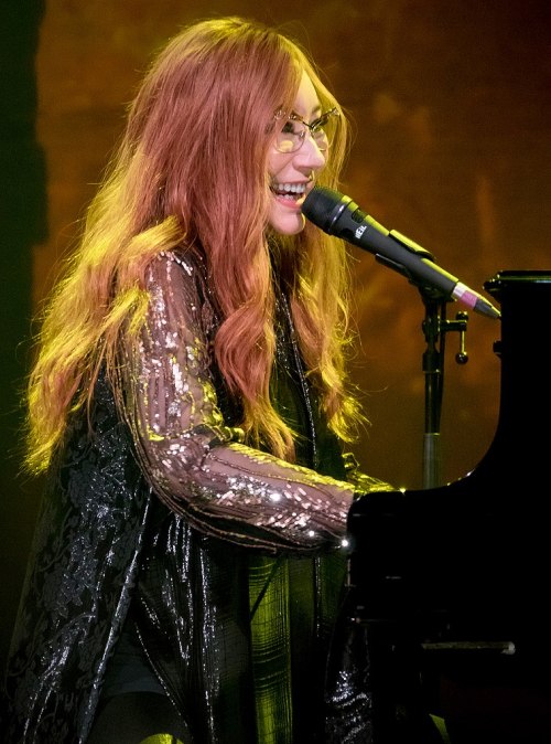A musician with long, wavy hair performs at a piano, smiling and singing into a microphone under warm stage lights.