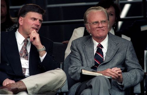 Two men seated together, one in a suit and tie, the other in a blazer, both looking thoughtfully at an event.
