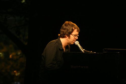A musician playing a piano and singing into a microphone on stage, with a dark background and soft lighting.