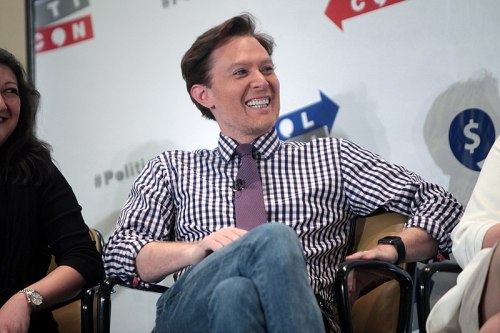 A smiling man in a checkered shirt and tie sits on a panel, engaging with the audience at an event.
