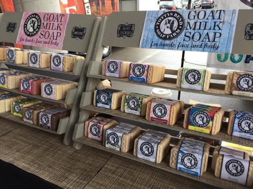 Shelves displaying various goat milk soaps in colorful packaging, labeled for hands, face, and body use.
