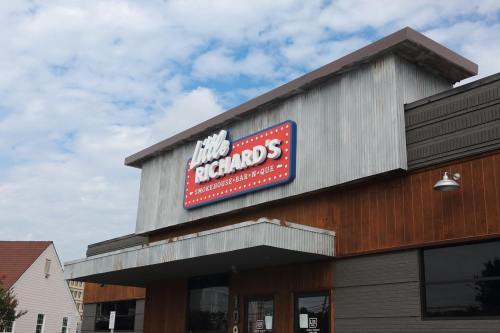 Sign for "Little Richard's Smokehouse Bar-B-Que" on a rustic building with a cloudy sky in the background.
